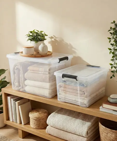 Zwei transparente Aufbewahrungsboxen mit gefalteten Textilien auf einem Holzregal, dekoriert mit Pflanzen und Körben.